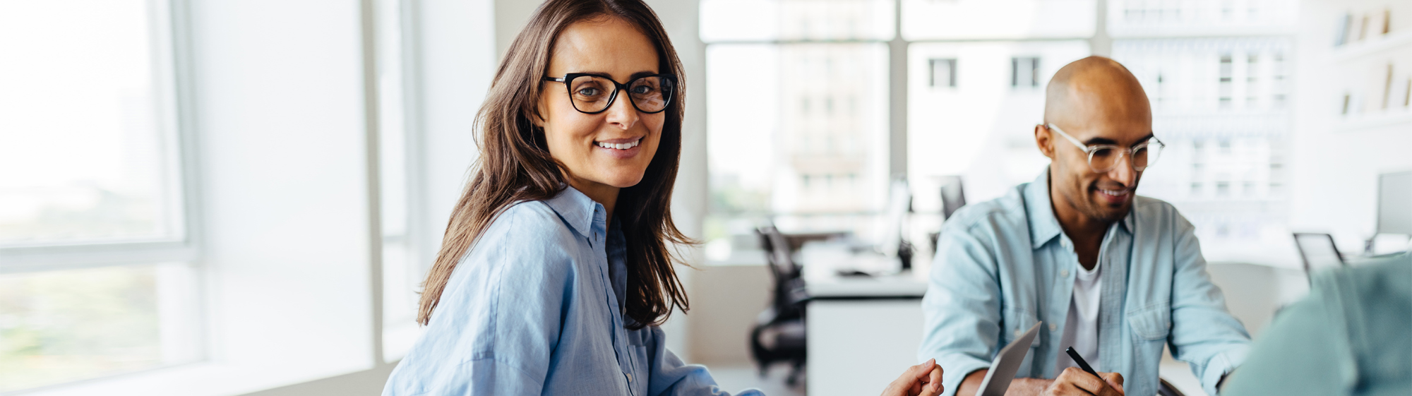 Eine Frau mit Brille im Büro dreht sich lächeln zur Kamera und ein Mann neben ihr ist lächelnd am Schreibtisch
