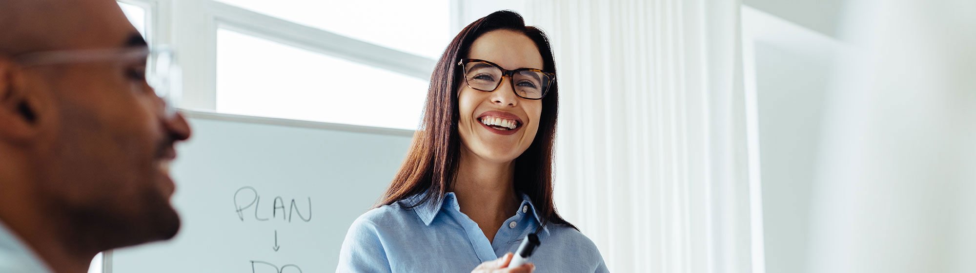 Ein Frau mit Brille lachend vor einem Flipchart mit Stift auf ihr Publikum zeigend