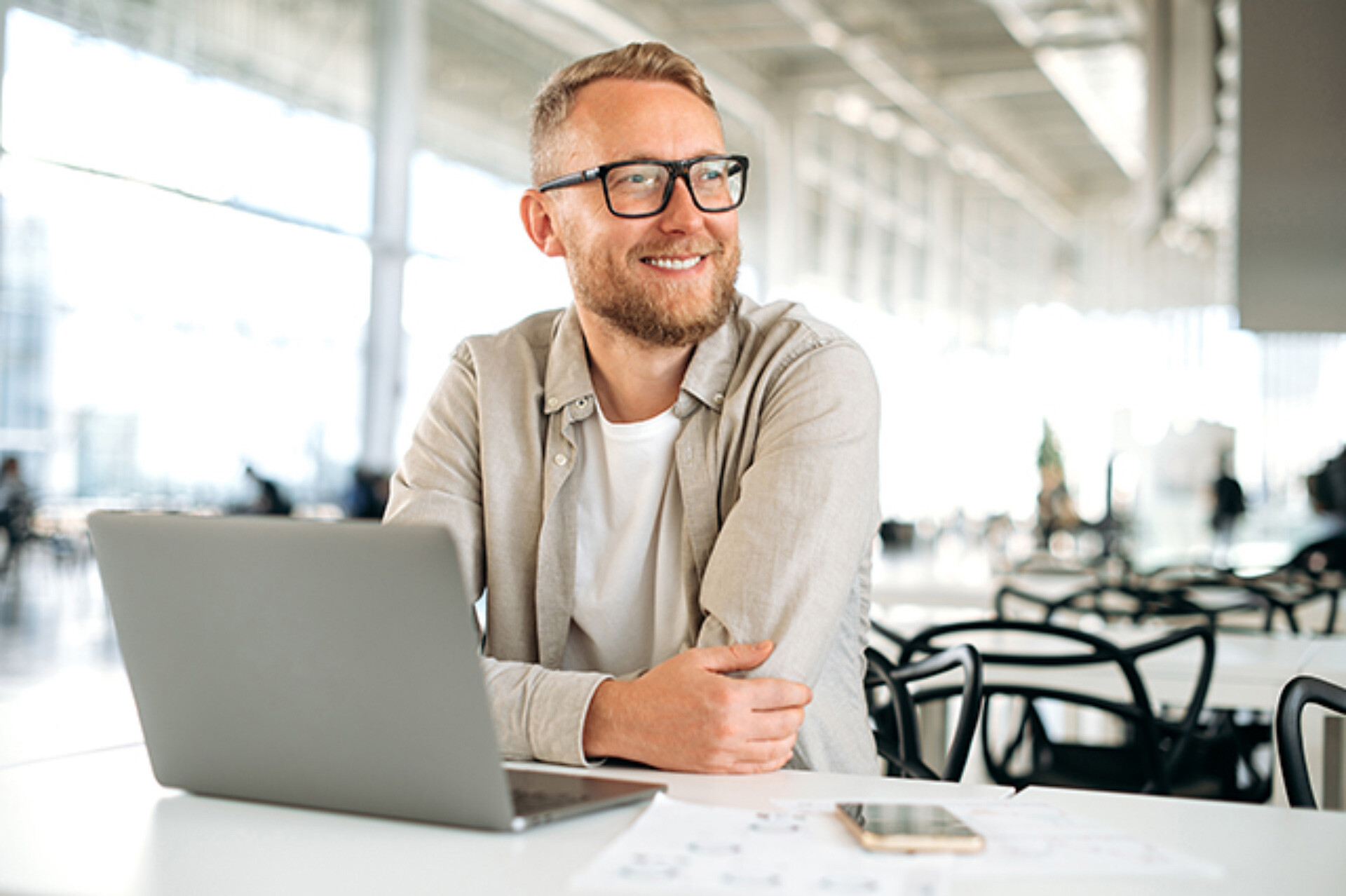 Ein Softwareentwickler sitzt entspannt an einem Schreibtisch mit Laptop und Unterlagen. In einer modernen, lichtdurchfluteten Umgebung denkt er über Lösungen nach und entwickelt innovative Software.